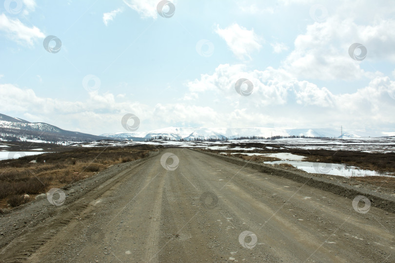 Скачать Грязная гравийная дорога пересекает огромную заснеженную поляну, окруженную высокими горными хребтами под пасмурным весенним небом. фотосток Ozero
