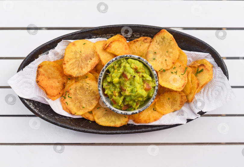 Скачать Домашние картофельные чипсы с вегетарианским соусом из авокадо и овощей на белом деревянном столе фотосток Ozero