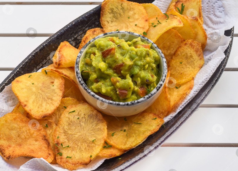 Скачать Домашние картофельные чипсы с вегетарианским соусом из авокадо и овощей на белом деревянном столе фотосток Ozero