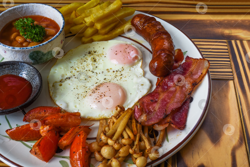 Скачать Полезный английский завтрак с яичницей-глазуньей, сосисками с беконом, картофелем фри, фасолью и помидорами крупным планом фотосток Ozero