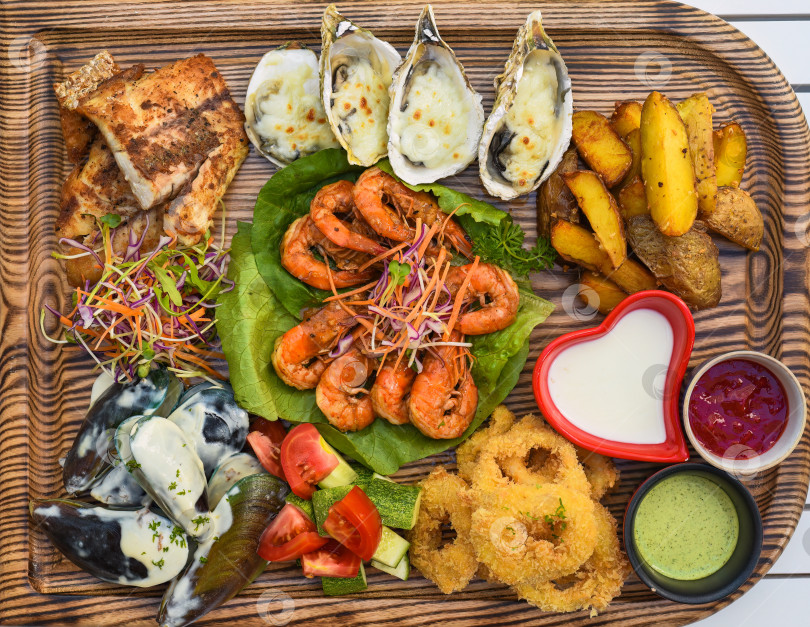 Скачать На столе много морепродуктов в виде жареной рыбы, кальмаров, устриц и соусов на деревянной доске фотосток Ozero