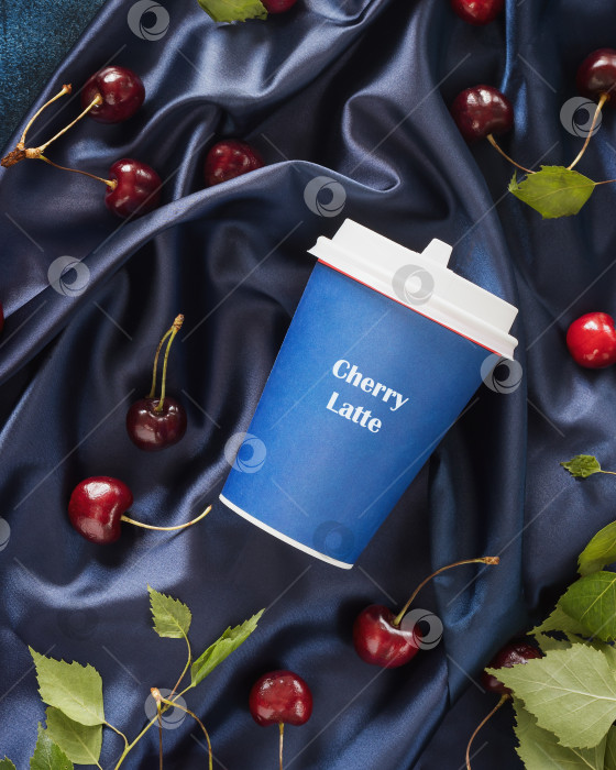 Скачать Кофейная композиция с крафтовой чашечкой вишневого латте, вишнями и листьями на голубом атласном фоне в складку фотосток Ozero