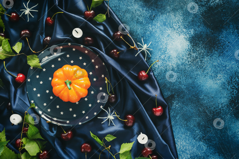 Скачать Тыква на тарелке, вишни, огарки свечей и декоративные пауки на голубой атласной ткани фотосток Ozero