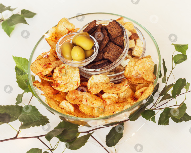 Скачать Ржаные крекеры, пшеничные крекеры, картофельные чипсы со специями и зелеными оливками в стеклянных вазочках на белом столе фотосток Ozero