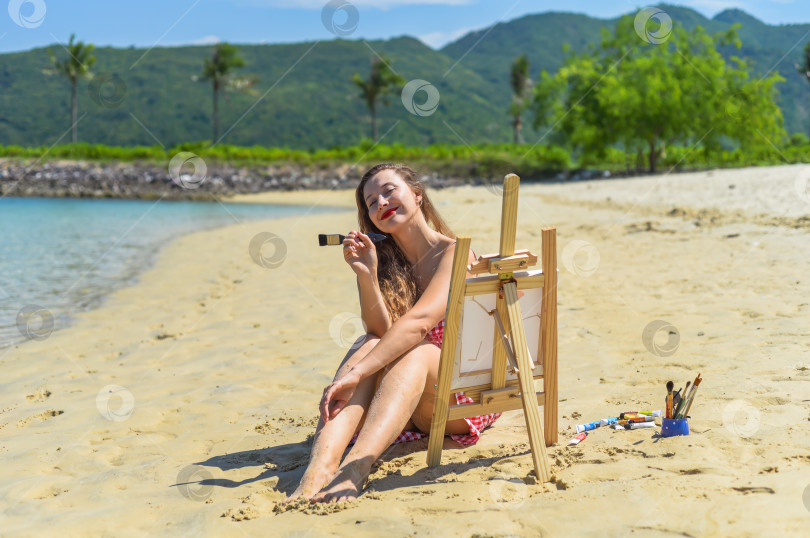 Скачать Молодая художница сидит летом на пляже с банкой красок, кистями и мольбертом рядом с морем и пальмами фотосток Ozero