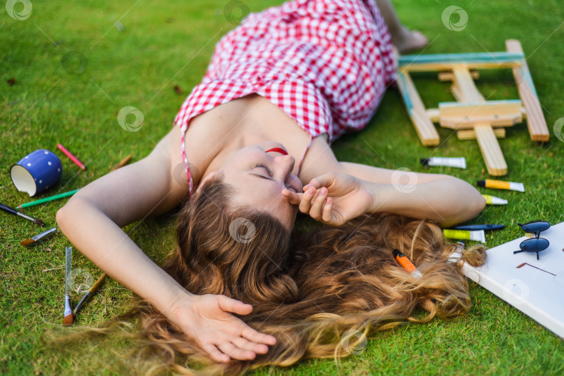 Скачать Красивая женщина, художница и иллюстратор, лежит на лужайке с брошенным мольбертом, рисунками, карандашами фотосток Ozero