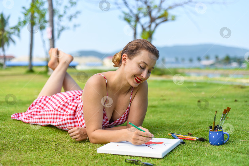 Скачать Молодая женщина, художница и иллюстратор, рисует карандашом, лежа летом на лужайке фотосток Ozero