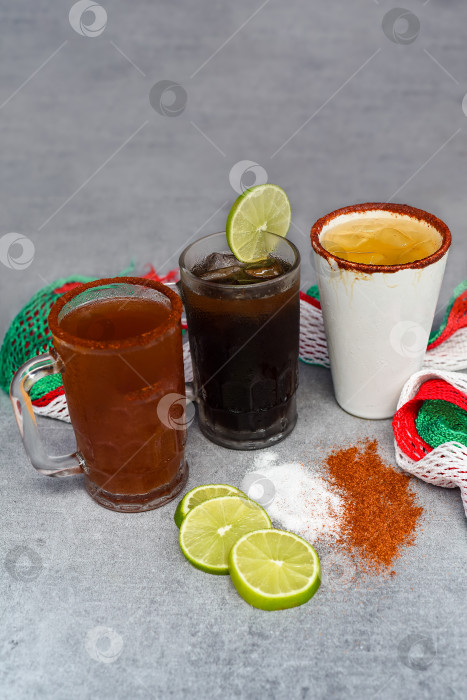 Скачать Мексиканские напитки "Чарро Негро", "Харрито", "Сервеза роха" подаются на сером столе с солью, перцем и лаймом фотосток Ozero