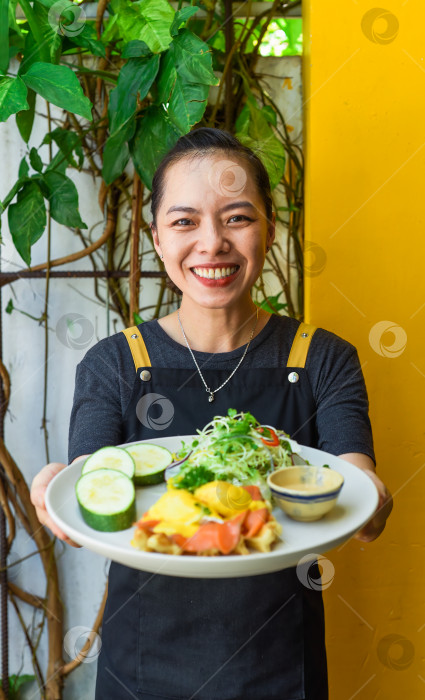 Скачать Молодая вьетнамская официантка держит тарелку с яйцами и вафлями Бенедикт с копченым лососем в ресторане фотосток Ozero