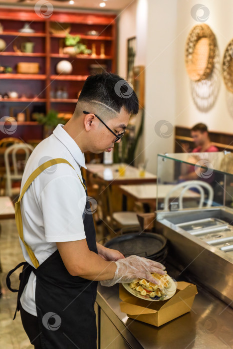 Скачать Вьетнамец-шеф-повар в униформе готовит свежеиспеченные бельгийские вафли с курицей и упаковывает их в бумажную коробку на вынос фотосток Ozero