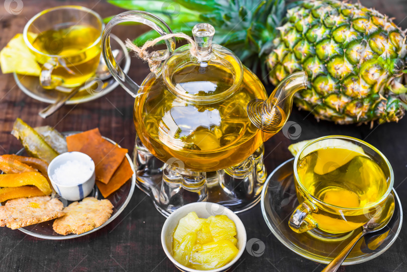 Скачать Стеклянный чайник с ананасовым чаем и домашним джемом, чашки и засахаренные фрукты на темном фоне фотосток Ozero