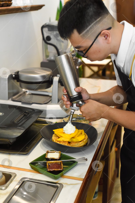 Скачать Шеф-повар-вьетнамец намазывает свежими сливками нарезанные фрукты манго на испеченные бельгийские вафли с помощью выжималки для сливок фотосток Ozero