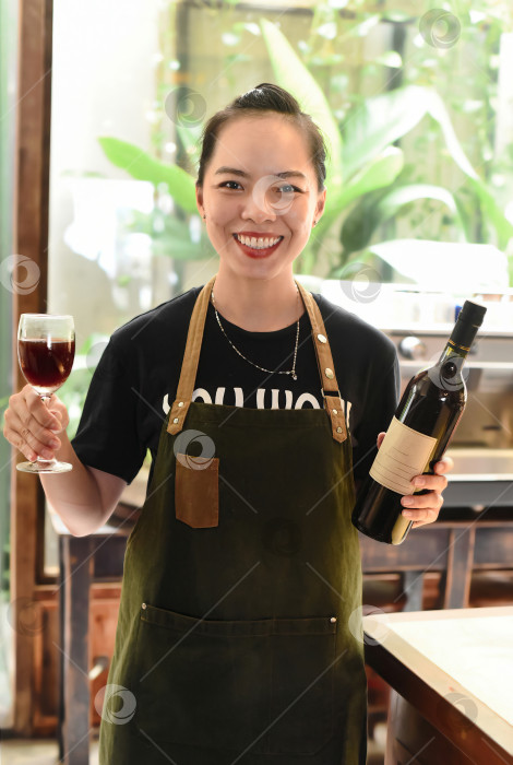 Скачать Счастливая вьетнамская официантка улыбается и держит в руках бутылку домашнего сливового вина и бокал вина фотосток Ozero