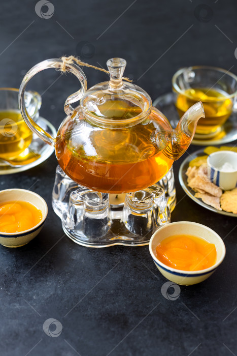 Скачать Стеклянный чайник со сливово-манговым чаем и ветчиной, чашки и засахаренные фрукты на черном фоне фотосток Ozero