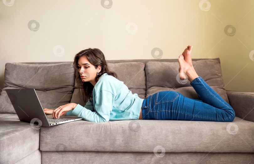 Скачать Молодая женщина лежит дома на диване и смотрит в ноутбук. Концепция онлайн-знакомств. она просматривает анкеты потенциальных партнеров в Интернете. Ноги скрещены под диваном фотосток Ozero