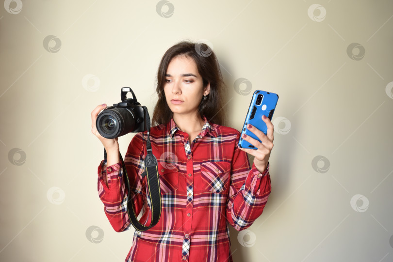 Скачать Молодая брюнетка-фотограф в красной рубашке, выбирающая между камерой и мобильным телефоном на бежевом фоне. Концепция выбора и разница между мобильной фотографией и камерой. Пространство для копирования фотосток Ozero