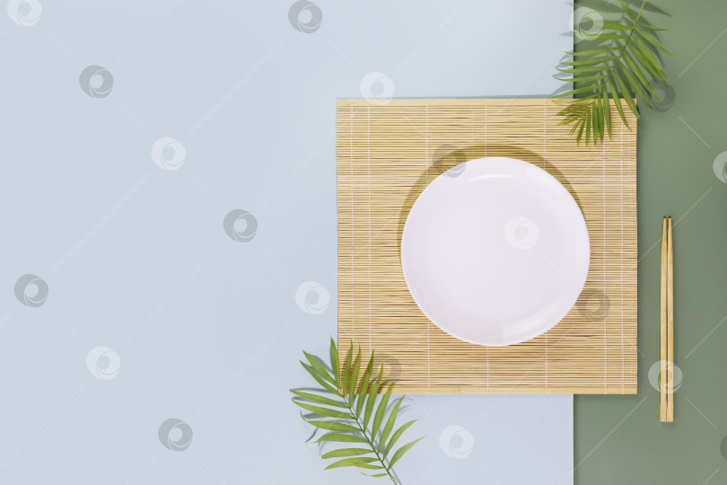 Скачать Пустая тарелка на природном коврике с деревянными палочками для еды и пальмовыми листьями фотосток Ozero