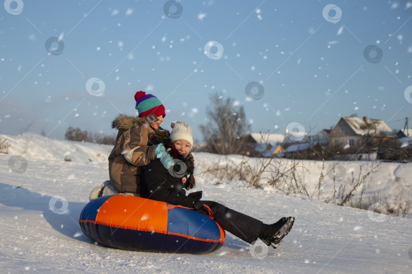 Скачать Зимним днем дети катаются на санках и весело смеются. Мальчик и девочка спускаются с горы на санках. Дети зимой в деревне на каникулах. фотосток Ozero