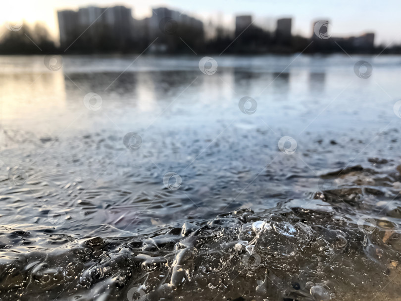 Скачать Замерзшая вода на краю озера с силуэтом многоэтажного здания на горизонте, зимний пейзаж фотосток Ozero