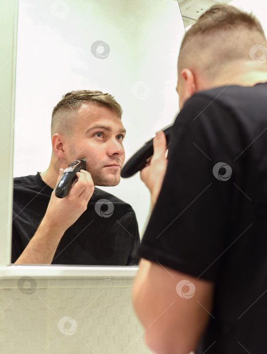 Скачать Молодой человек бреется триммером и смотрится в зеркало в ванной комнате фотосток Ozero