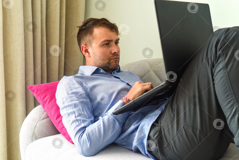 Скачать Молодой человек-фрилансер, работающий дома со своим ноутбуком, лежа на диване фотосток Ozero