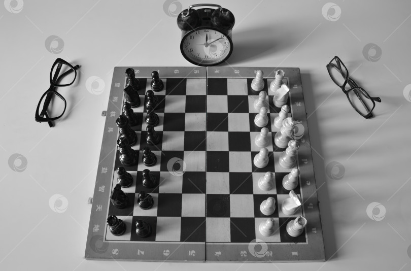 Скачать Черно-белая фотография старой шахматной доски с изъянами и фигурами. По бокам очки, потертый пыльный будильник показывает время. фотосток Ozero