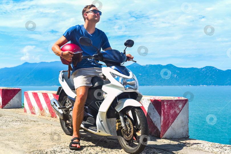 Скачать Молодой человек, водитель мотоцикла, сидит и держит в руках шлем на фоне южно-Китайского моря во Вьетнаме фотосток Ozero