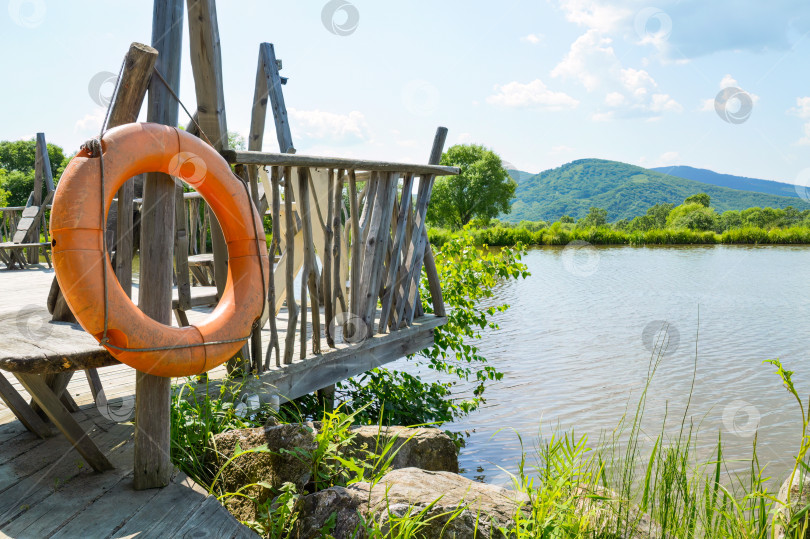 Скачать Оранжевый спасательный круг, висящий на пирсе у пруда в солнечный летний день фотосток Ozero