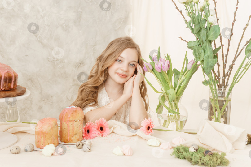 Скачать Девушка с длинными волосами в светлом платье сидит за пасхальным столом с куличами, весенними цветами и перепелиными яйцами. Счастливого празднования Пасхи. фотосток Ozero