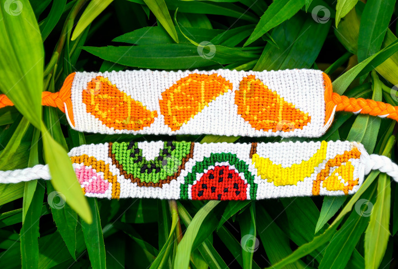 Скачать Два плетеных браслета дружбы ручной работы из ниток с фруктовыми узорами на фоне зеленых листьев фотосток Ozero