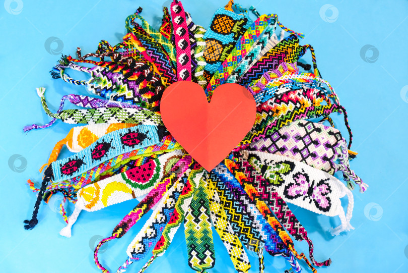 Скачать Красное сердечко в центре круга из множества разноцветных плетеных браслетов дружбы ручной работы, вышитых яркой нитью с узелками на синем фоне. фотосток Ozero
