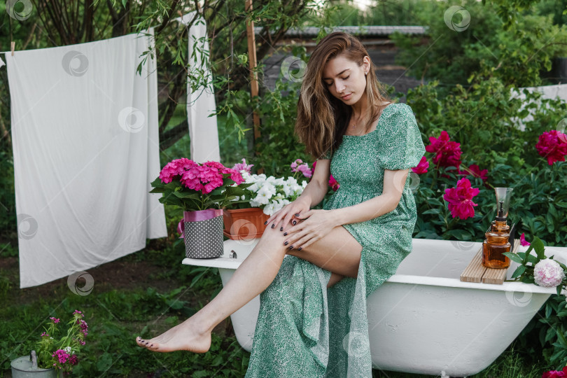 Скачать Женщина сидит на чугунной ванне во дворе загородного дома рядом с кустом цветущих пионов. Концепция лета, загородной жизни, ванной комнаты на улице в цветущем саду за городом фотосток Ozero