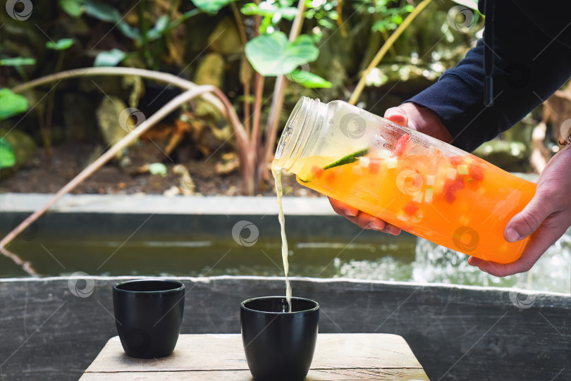 Скачать Руки держат бутылку со свежим домашним соком из апельсинов, арбуза, лайма и наливают в чашку фотосток Ozero