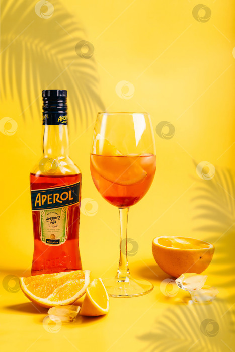 Скачать Бутылка летнего коктейльного напитка Aperol Aperitivo с бокалом для коктейля spritz и апельсинами на ярко-желтом фоне фотосток Ozero