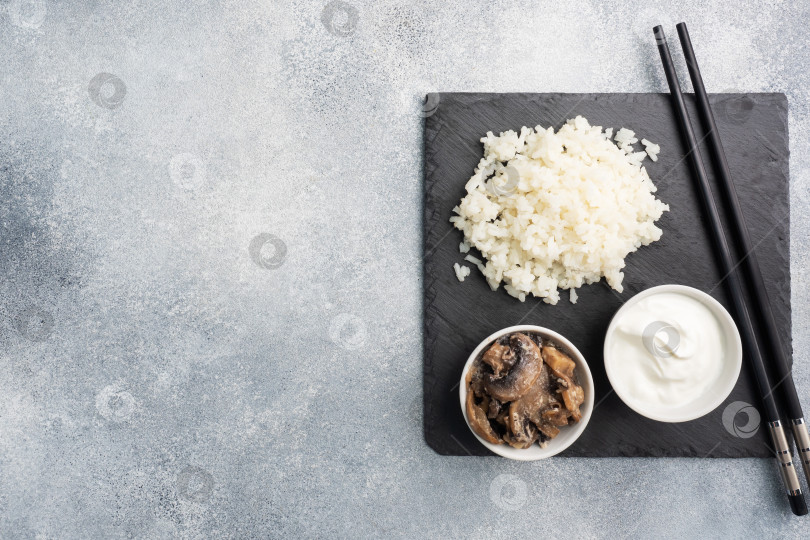 Скачать Отварной белый рис и грибы в сметане на черной подставке с палочками для еды. Серый бетонный фон копирует пространство. фотосток Ozero