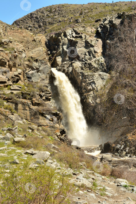 Скачать Бурный поток водопада стремительным потоком вытекает из расщелины в горах солнечным весенним днем. фотосток Ozero