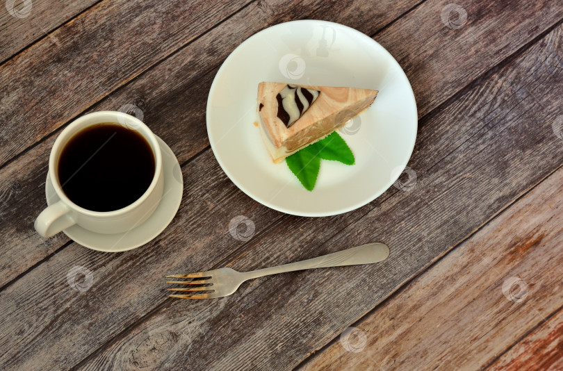 Скачать Чашка горячего кофе на блюдце, вилка и тарелка с кусочком свежего сливочного чизкейка с листьями мяты на столе из темного дерева. фотосток Ozero