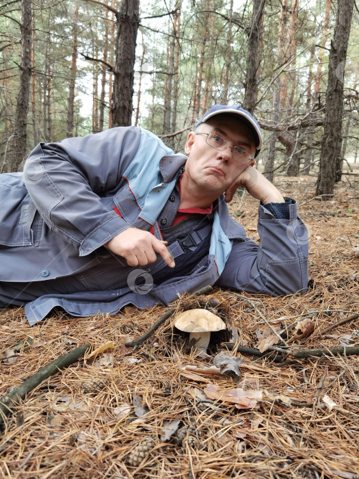 Скачать мужчина в комбинезоне очках и кепке лежит на иголках в сосновом лесу и указывает пальцем на подберезовик фотосток Ozero