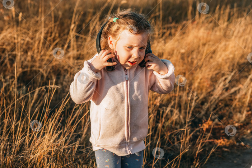 Скачать Маленькая девочка слушает музыку в наушниках, портрет забавного малыша с голубыми глазами крупным планом, ребенок на улице в солнечный осенний день. фотосток Ozero