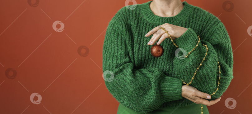 Скачать Женщина в зеленом свитере держит в руках символ Нового года - красный елочный шар. имитация рождественской елки фотосток Ozero