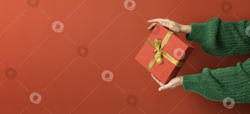 Скачать Женские руки преподносят рождественскую красную подарочную коробку с золотым бантом на красном фоне. Зеленый свитер. Концепция Нового года. Зимние праздники и подарки. Доставка сюрпризов фотосток Ozero