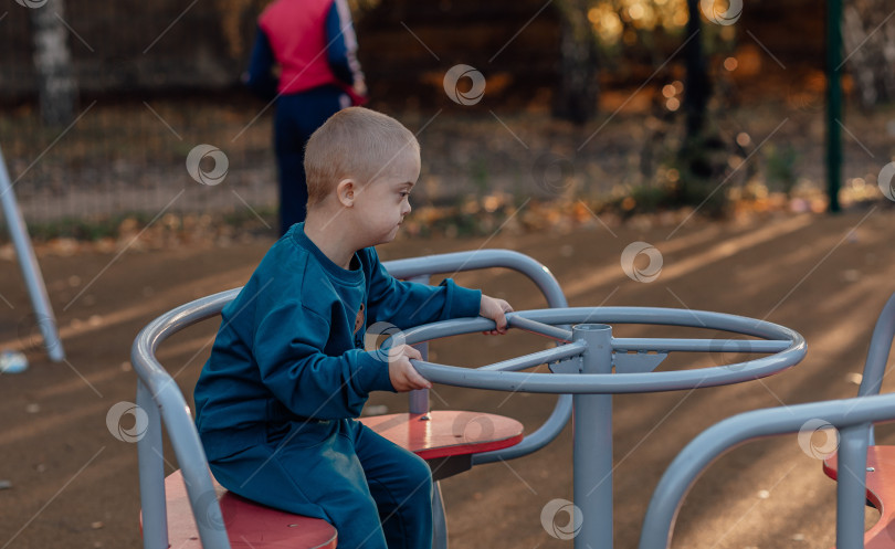 Скачать Милый маленький мальчик с синдромом Дауна в забавной шапочке гуляет по детской площадке, крутится на карусели фотосток Ozero
