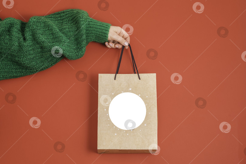 Скачать Концепция рождественских покупок. Женская рука в зеленом свитере, держащая подарочный пакет на красном фоне, фотосток Ozero