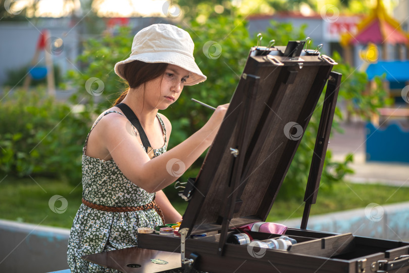 Скачать Симпатичная девочка-подросток рисует за мольбертом на открытом воздухе, сделай сам фотосток Ozero