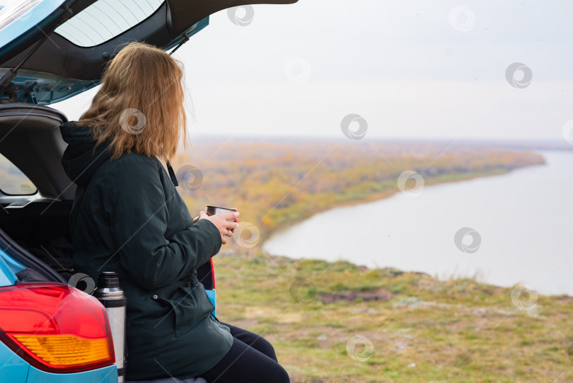 Скачать Счастливая молодая белая женщина в свитере пьет чай, сидя в багажнике автомобиля на высоком берегу реки фотосток Ozero
