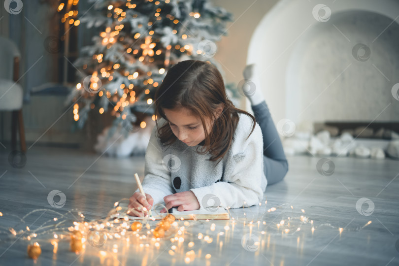 Скачать симпатичная девочка лежит на полу возле рождественской елки и пишет письмо Деду Морозу фотосток Ozero