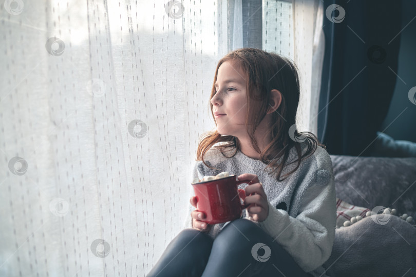 Скачать симпатичная девушка на подоконнике держит красную кружку с какао и зефиром и смотрит в окно. фотосток Ozero