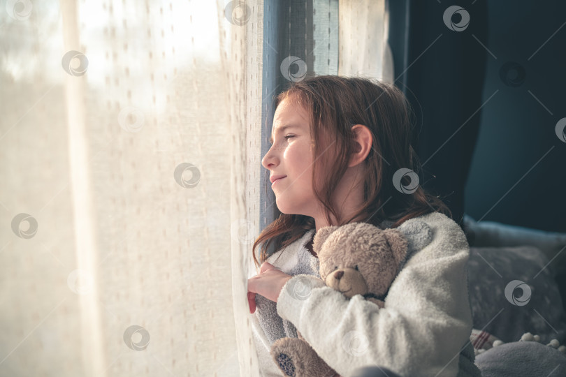 Скачать задумчивая милая девушка сидит и смотрит в окно, обнимая плюшевого мишку. фотосток Ozero