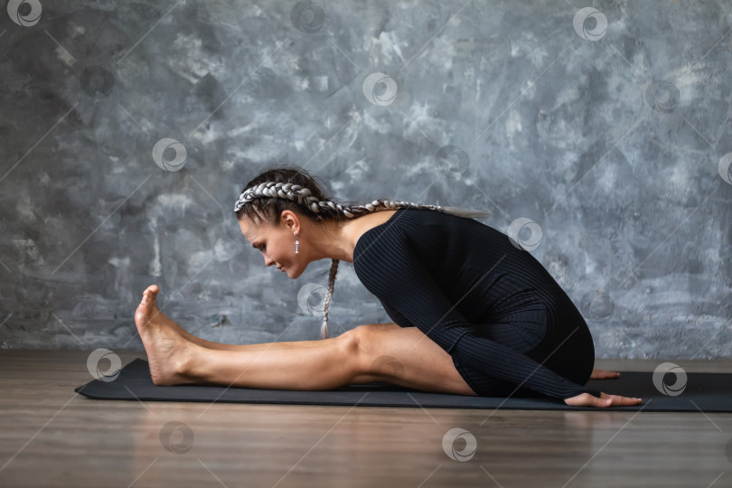 Скачать Женщина средних лет выполняет упражнение для растяжки мышц спины и ног, наклоняется вперед из положения сидя, тренируется в спортивной одежде на коврике у стены фотосток Ozero