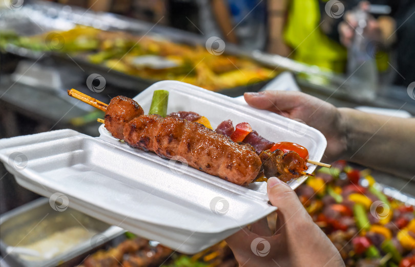 Скачать Руки держат пластиковую коробку с шашлыком на шпажках на вьетнамском ночном рынке во время фестиваля еды фотосток Ozero